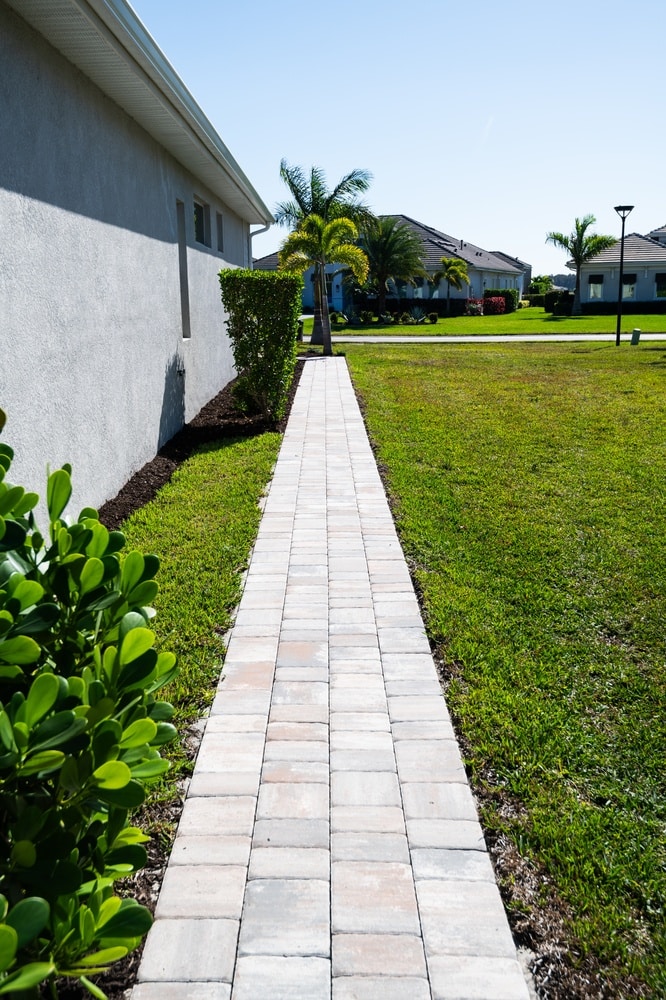 Allée en Pavés pour Aménagement Extérieur Moderne Allée en pavés autobloquants longeant une maison moderne avec pelouse verte et palmiers sous un ciel bleu.