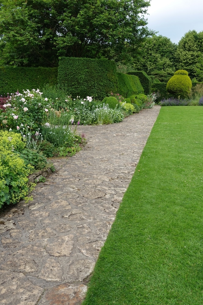 Jardin paysager avec allée en pierre et bordures fleuries Allée en pierre naturelle bordée de fleurs, pelouse verte et haies de jardin sculptées avec soin.