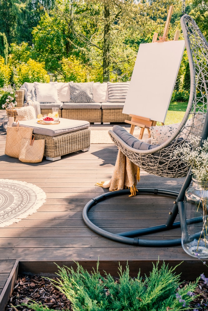 Déco terrasse jardin : canapé osier et fauteuil suspendu Terrasse en bois avec canapé en osier, fauteuil suspendu et chevalet blanc dans un jardin ensoleillé et verdoyant.