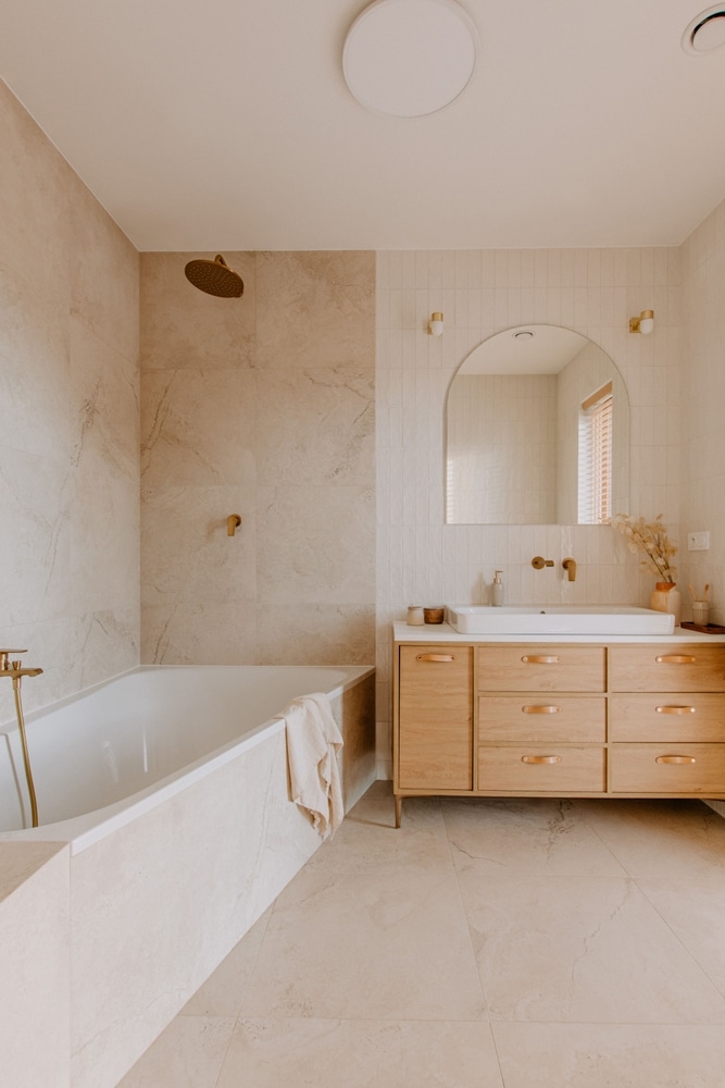 Salle de bain moderne beige avec bois et touches dorées Salle de bain moderne avec baignoire, carrelage beige, robinetterie dorée et meuble sous vasque en bois.
